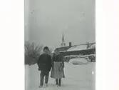 Två barn står i snön, flickan håller i böcker. I bakgrunden står ett timrat hus med vedtravar och en eka framför. Mariefreds kyrkspira sticker upp över taket. (Samma motiv som E5418)