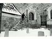 Snöbild. En man i rock och hög svart hatt går nedför en trappa från en stor gammal stenkyrka. Till höger i bild syns en gravsten över grosshandlare H L Broman med maka J C Ljungdahl.