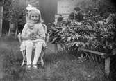 Ruben Liljefors dotter Marit sitter på stolen i trädgården vid pionbuske med sin docka i famnen, sannolikt Svensgården i Dalarna