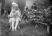 Ruben Liljefors dotter Marit sitter på stolen i trädgården vid pionbuske med sin docka i famnen, sannolikt Svensgården i Dalarna