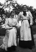 Två glada kvinnor, en sitter och den andra står, ute i trädgård, sannolikt i Sverige
