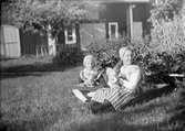 Två flickor, den ena Marit Liljefors, klädda i folkdräkt, sitter på gräsmattan ute i trädgård, sannolikt Svensgården, Dalarna