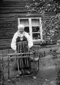 Gammal kvinna, klädd i folkdräkt, står framför huset bakom härvel, sannolikt Dalarna