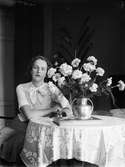Ung kvinna sitter vid bord med blommor 1930-40-tal, sannolikt i hemmet i Sverige
