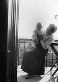 Kvinna sitter på balkong med liten flicka i famnen, sannolikt i Sverige