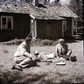 Kloar-Erik och Anders Lundgren sitter i gröngräset en stuga med sticktak/pärt, Dämmarehult, med kaffepanna på ett fotogenkök.