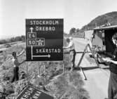 Nya skyltar sätts upp på E4:an vid norra utfarten från Huskvarna. Mannen till höger med penna och block är Arne Putzén som vid den tiden var tidningen Smålands Folkblads lokalreporter. 1960-tal.