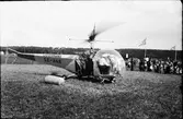 En helikopter som står på backen med en folkskara i bakgrunden. På helikoptern står det Österman SE-HAE och två män syns i cockpiten.