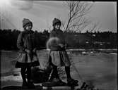 Två flickor står på kälkar på isen i solsken, iklädda mössor, sjalar, stickade tröjor, kjolar och träskor men barhänta.