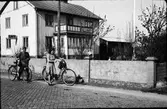 Kvinna och militär med cyklar passerar ett Café Hembageri, möjligen Bengtssons bageri i Åskloster. Tomten omgärdas av en putsad mur med lejonmaskaroner och kantställda kuber på pelarna.