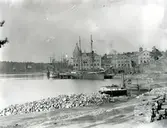 Vy från Kattholmen mot hamnen med skonare samt ångbåt förtöjda vid kajen. Till vänster syns Huvudkontoret uppfört 1898, samt de båda packbodarna. Statbyggningen och Ekensberg skymtar till vänster i bakgrunden. De sistnämnda bygganderna revs när Gustavsbergs kyrka stod färdig 1907.