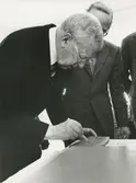 Interiör. Kungligt besök på Gustavsbergs Fabriker. Stig Lindberg,och Kung Gustav VI Adolf. Kungen skriver sin signatur i lera. Sannolikt på Keramiskt Centrum.
Personer: Stig Lindberg och Kung Gustav VI Adolf