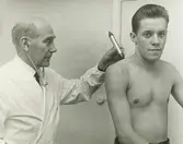 Interiör. Sjukrummet, företagshälsovård på Gustavsbergs Fabriker.. Läkarundersökning av anställd.
Personer: Doktor Bellander och okänd