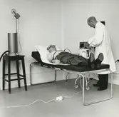 Interiör. Sjukrummet, företagshälsovård på Gustavsbergs Fabriker.. Läkarundersökning av anställd.
Personer: okänd och Doktor Bellander