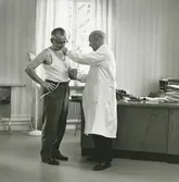 Interiör. Sjukrummet, företagshälsovård på Gustavsbergs Fabriker.. Läkarundersökning av anställd.
Personer: Doktor Bellander och Gösta Dahlberg, personalchef