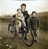 Jenny Lidberg på promenad med cykeln och barnen. Lilla Elly sitter på en extra sits monterad bakom styret och Leif går bredvid. De är klädda för en kylig dag med öronlappsmössor, varma jackor och vantar.