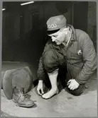 Personvagnsreparatör Einar Larsson, vid Trafikaktiebolaget Grängesberg–Oxelösunds Järnvägars järnvägsverkstad i Eskilstuna, demonstrerar hur skyddsskon räddade hans tår i samband med en arbetsplatsolycka då en 150 kilos buffert föll ner på hans fot.