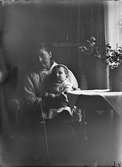 I hemmiljö där en äldre kvinna sitter med ett litet barn i knäet, troligtvis 1920-30-tal.