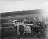 Studenter, kvinnor och män sittande vid bord ute på gräs, skog i bakgrunden, sannolikt Uppland omkring 1900