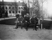 Tre män iklädda kostym och studentmössor sittande på bänk i Universitetsparken, Uppsala omkring 1900