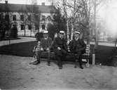 Tre män iklädda kostym och studentmössor sittande på bänk i Universitetsparken, Uppsala omkring 1900