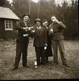 Tre män och en kvinna poserar finklädda utanför Kloar-Eriks stuga med kaffekoppar och spritflaska, kaffegök således. Kvinnan håller en liten bukett med påskliljor så det är vår. På marken framför dem står en kaffetermos. Från vänster: Målar-Anders från Röräng, 