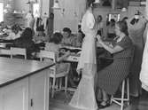 Interiör från NK, NK:s Franska damskrädderi, ateljén. Runt ett stort bord sitter sömmerskor, några kvinnor syr, en uppsätterska modellerar en toall på en provdocka (mannequin). 26 augusti 1943.