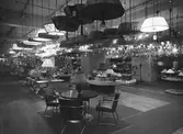 Interiör från varuhuset NK i Stockholm. På lampavdelningen hänger takarmaturerna i långa rader. Bordslampor exponeras på hyllor och bord. I förgrunden en sittgrupp med stålrörsmöbler.