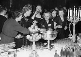 En grupp festklädda kvinnor tar för sig ur en bålskål vid ett elegant dukat bord. Man firar Nordiska kompaniets 50-årsjubileum i januari 1952. Lokalen är sannolikt Stockholms stadshus.