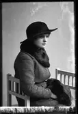 Porträtt av Vivan Hedlund sittandes på en pall och klädd i svart bredbrättad hatt, ytterkappa samt krage och muff i mörk päls.