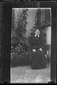 Reprofotografi av bild på en svartklädd kvinna som sitter utanför ett hus. Bakom henne syns en hund och en blomsterrabatt.