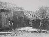 Ryggåsstuga med s k skunke vid entrén. Framför huset är marken belagd med stenhallar och där står en man i väst och keps. Bilden märkt 