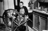 Äldre kvinna, Amanda Dahlgren, sitter vid spinnrocken på Kungsgatan 8 och spinner. Hon är klädd i kofta och förkläde.
