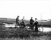 Gruppbild med tre kvinnor i kjol och hatt samt en pojke i sjömansskjorta som står på ett öppet fält med var sin cykel. I bakgrunden syns en sjö och en landsväg. Avbildade: Annie Michelsen, Maria Eriksson, Nella Hedén och Anders Hedén.