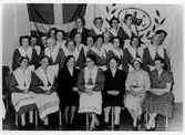 Gruppbild av 26 kvinnor från Alingsås Kooperativa Kvinnogille varav 19 står och sju är sittande. I bakgrunden ses svenska flaggan samt en organisationsflagga. Bilden tag under deras 25-årsjubileum 1933-1958.