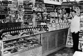 Inifrån Gunnar Johanssons speceriaffär. En man står framför disken i en vit skjorta. Väggarna och disken är fyllda med diverse varor.
Kvarteret Storken.