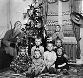 Olof Jacobsen, utklädd till tomte, sitter vid en julgran tillsammans med en liten skara barn hos familjen Bolin, Nolby. Olof (Olle) Jacobsen var far till Monica Jacobsen (andra från vänster i främre raden) och Dick Jacobsen (pojken längst bak).