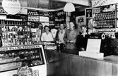 Inifrån Sandbergs speceriaffär. Bakom disken står 3 män, 1 kvinna och 1 tonårspojke. Till vänster en stor glasdisk fylld med olika sillinläggningar.
Kvarteret Storken.
