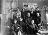 Gruppbild med delar av fräslningsarmén, 7 kvinnor och 4 män, hemma hos en av medlemmarna, Hilma Andersson, Annelund, Dammen. Bakom dom ses ett piano, ett fönster, en väggbonad och ett blomsterstativ.