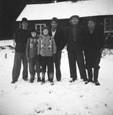 Två pojkar med skridskoskenor om halsen står omgivna av fyra män i snön utanför en stuga, troligen i Börsgård då en person anges vara Åke i Börsgård. Längs till höger står Viktor på Storåker och bredvid honom 