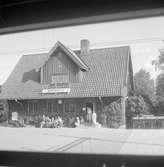 Tystberga station med en samling män och kvinnor utanför stationshuset. Fotot ser ut att vara taget från en innestående tågvagn.