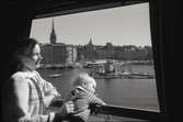 Kvinna med barn som tittar ut genom kupé fönster under resa på Södra Järnvägsbron, på sträckan mellan Riddarholmen och Mälarstrand. Tyska Kyrkans torn i bildens vänstra del.