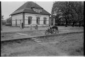 Stationshuset i Åsbo-Össjö. Tågtrafiken upphörde 1953-06-10. Barn leker med motorcykel på plattformen. Spåren är borttagna.