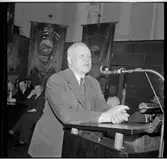 Man som talar i mikrofon vid en talarstol. I bakgrunden ses standar.