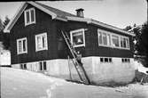 Familjen Lundgrens hus i Ry, Veddige. Fasaden har en ovanlig stockliknande panel och kanske har man just satt in perspektivfönstren (de vändbara introducerades 1943, dessa är icke vändbara) på verandan. En stege står lutad mot fasaden och dottern Ella sitter halvvägs upp på den.