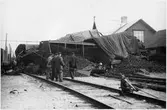 Arbete på urspårade godsvagnar lastade med tackjärn samt med stationshus i bakgrunden.