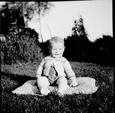 Lilla pojken Roy sitter på en dyna i gräset. Han har kofta, strumpor och skor till sin byxdress.