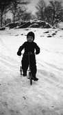 Ett litet barn med trehjuling i snön, klädd i overall med skärp, vantar, mössa och kängor.