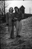 Två finklädda flickor, Iris t.v. och Mary t.h, i vida 1940-talsbyxor, hattar och broscher står arm i arm i en backe med lastbilar och en elcentral i bakgrunden  (se även foto EA1183).