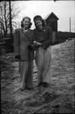 Två finklädda flickor, Iris t.v. och Mary t.h, i vida 1940-talsbyxor, hattar och broscher står arm i arm i en backe med lastbilar och en elcentral i bakgrunden  (se även foto EA1183).
